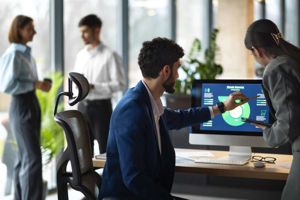 Business colleagues collaborating in a modern office, analyzing a circular economy chart on a computer. This scene illustrates the integration of ERP and CRM systems for data-driven strategic planning and promoting sustainable business practices.