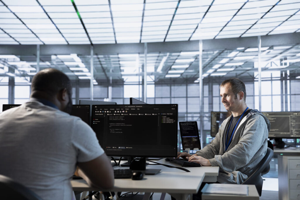Two professionals working at an IT service desk in a modern office, using multiple monitors that display lines of code to manage and resolve technical issues.
