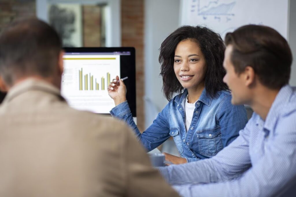 A team of professionals collaborates in a meeting, with a woman pointing to a bar chart on a computer screen to explain insights derived from Salesforce CRM Analytics.