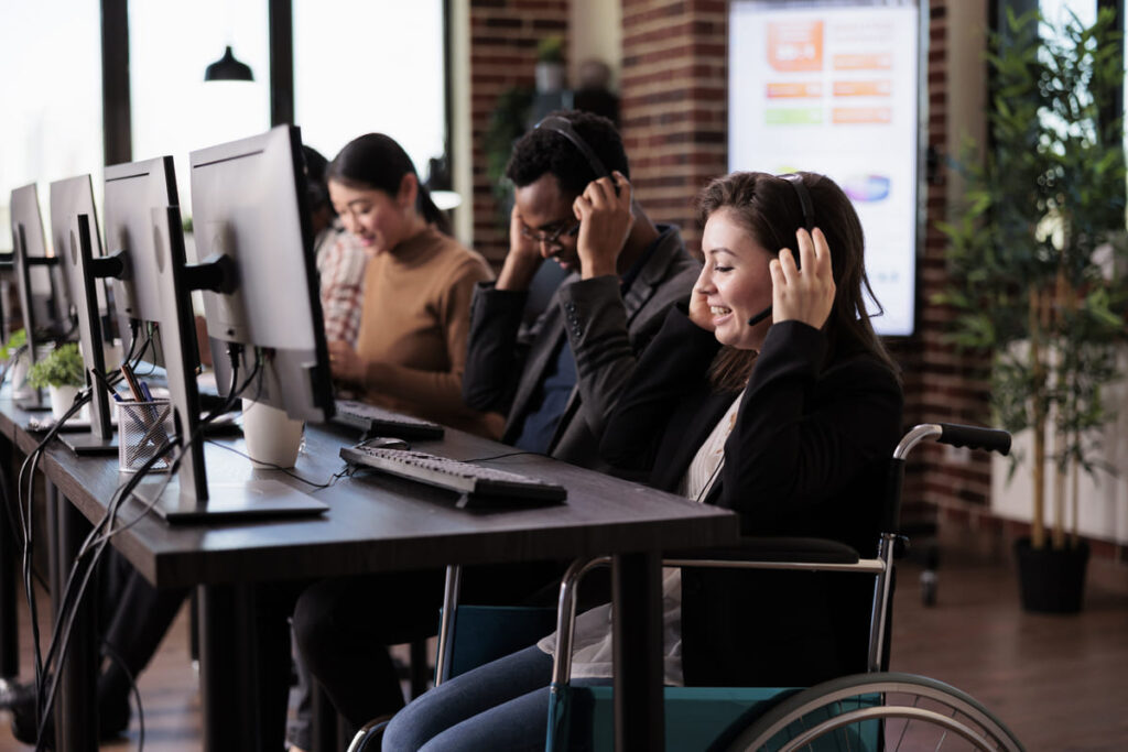 Customer support professionals working at computer stations, including a woman using a wheelchair, illustrating an accessible work environment aligned with principles of inclusive design.