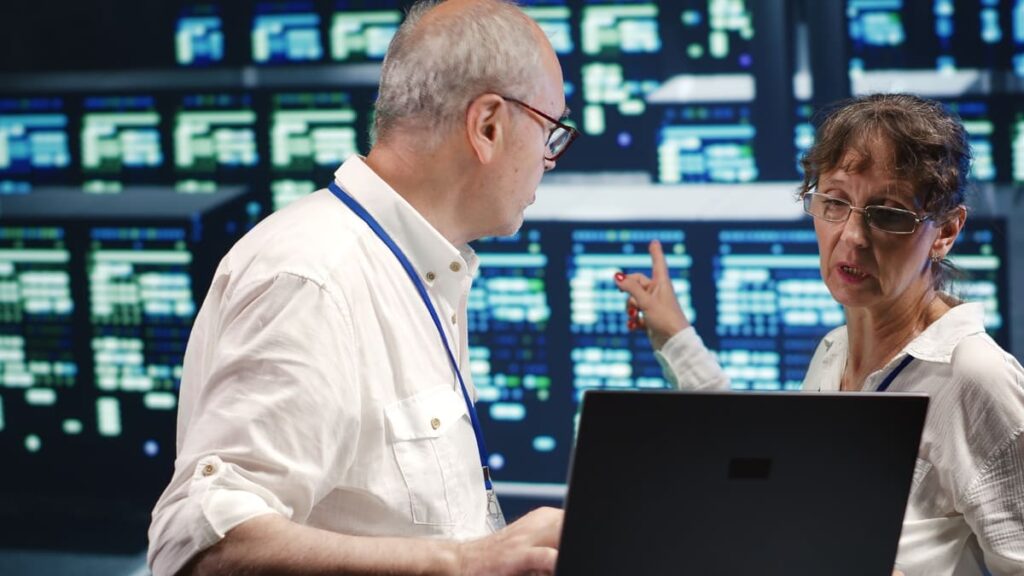 Two IT professionals analyze data on a laptop in front of server screens, discussing strategies for Cloud Risk Management.