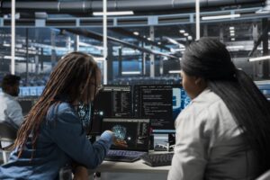 wo female engineers collaborating in a modern tech office with server racks in the background, analyzing code and network visualizations on a laptop, representing the management of Infrastructure as Code.