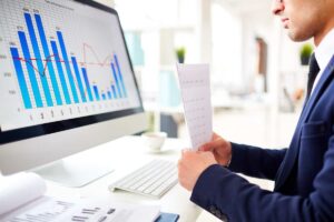 Side profile of a financial professional in a suit at a modern desk, carefully reviewing a printed document while analyzing complex bar and line charts on a large computer monitor, representing the critical role of data management in financial services.